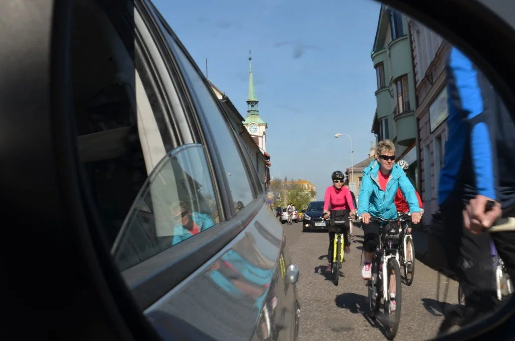 Cyklisté v zrcátku auta projíždějí ulicí, v pozadí věž