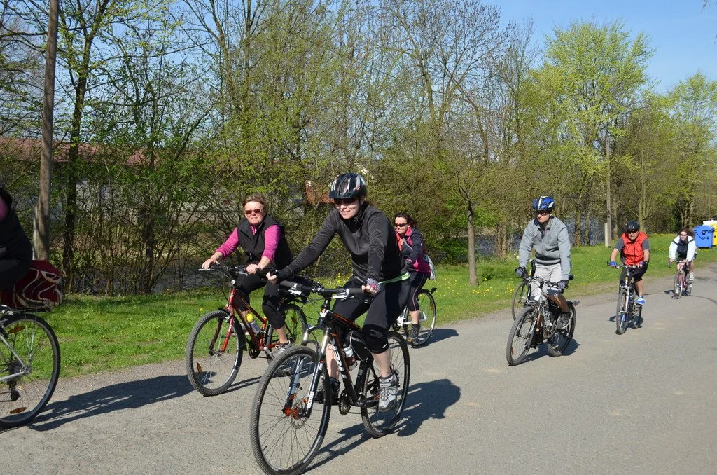 Skupina cyklistů projíždí alejí podél řeky na jarní vyjížďce