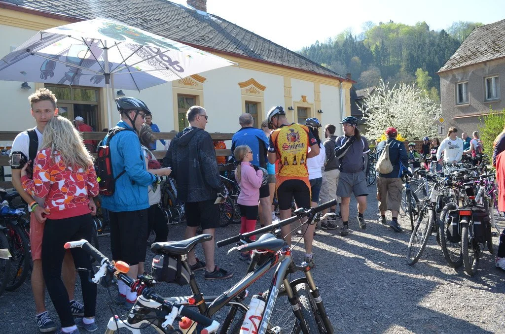 Cyklisté odpočívají pod slunečníkem před budovou u zalesněného kopce