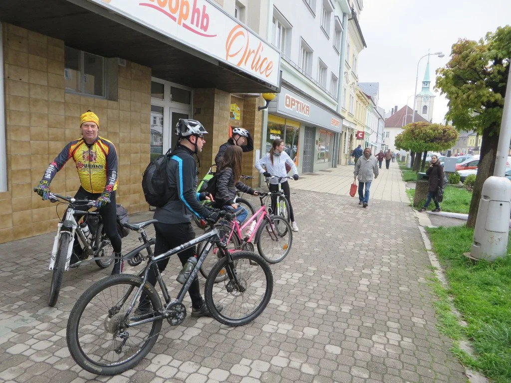 Cyklisté s koly na pěší zóně před obchody, v pozadí kostelní věž