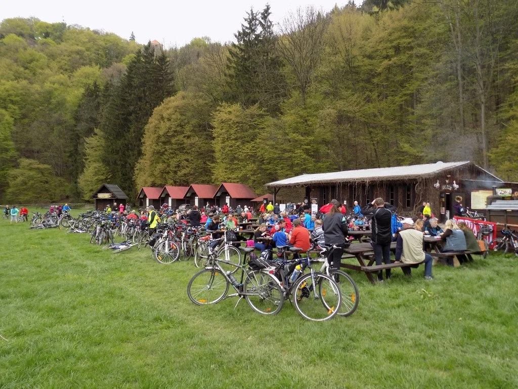 Cyklisté vedou kola kolem dřevěné chaty s věžičkou a karavany