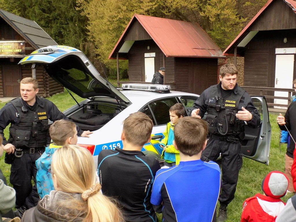 Policista otevírá kufr vozu před skupinou dětí u chatek v lese