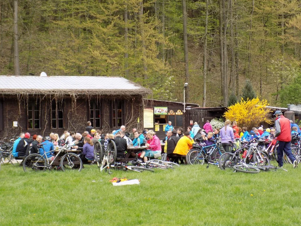 Velká skupina cyklistů u dřevěné chaty v lese s množstvím zaparkovaných kol