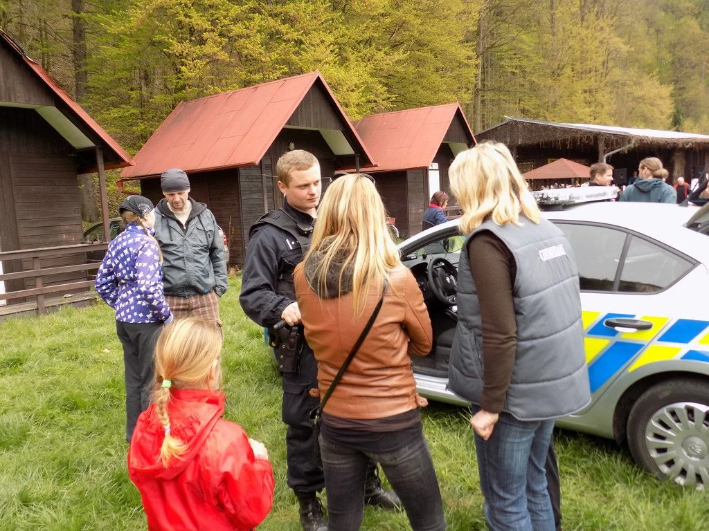 Lidé a děti u policejního vozu mezi dřevěnými chatkami