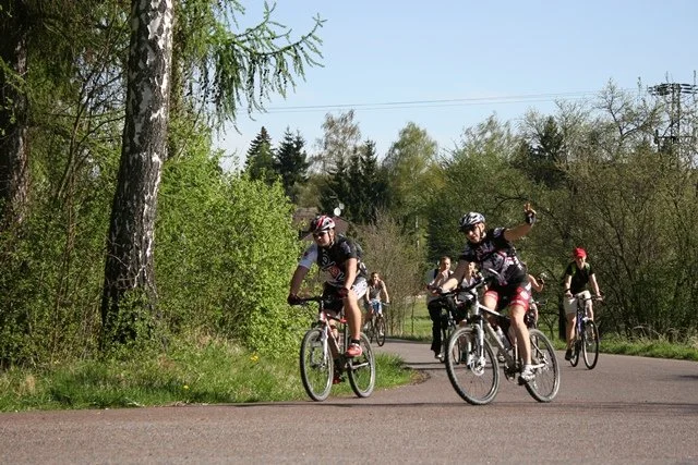 Dva cyklisté v dresech jedou po cestě mezi zelení, jeden mává