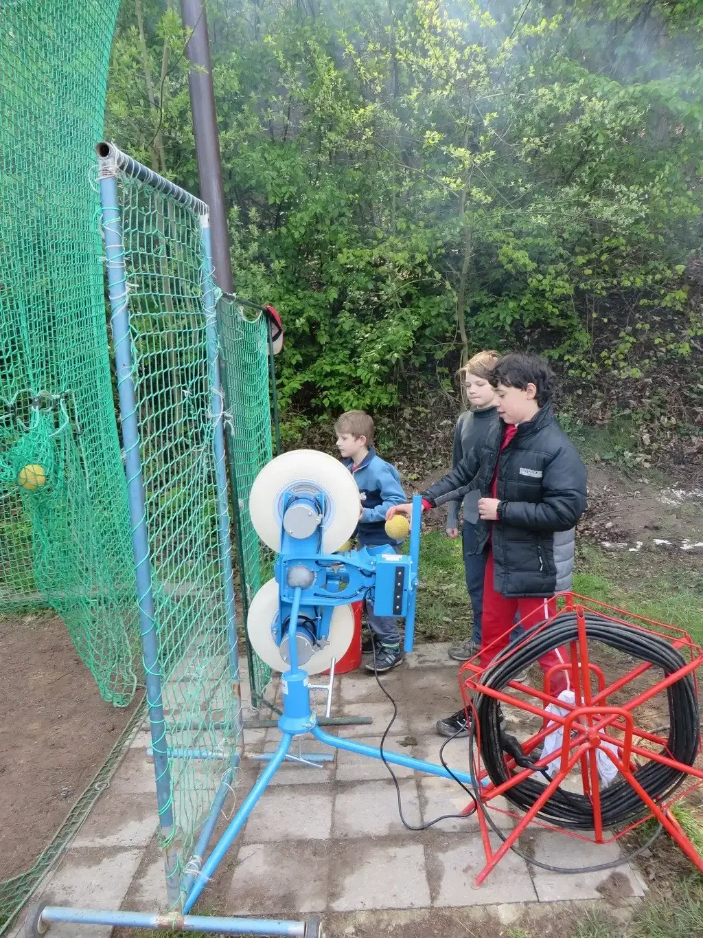 Děti obsluhují míčkovací stroj u sportovní sítě