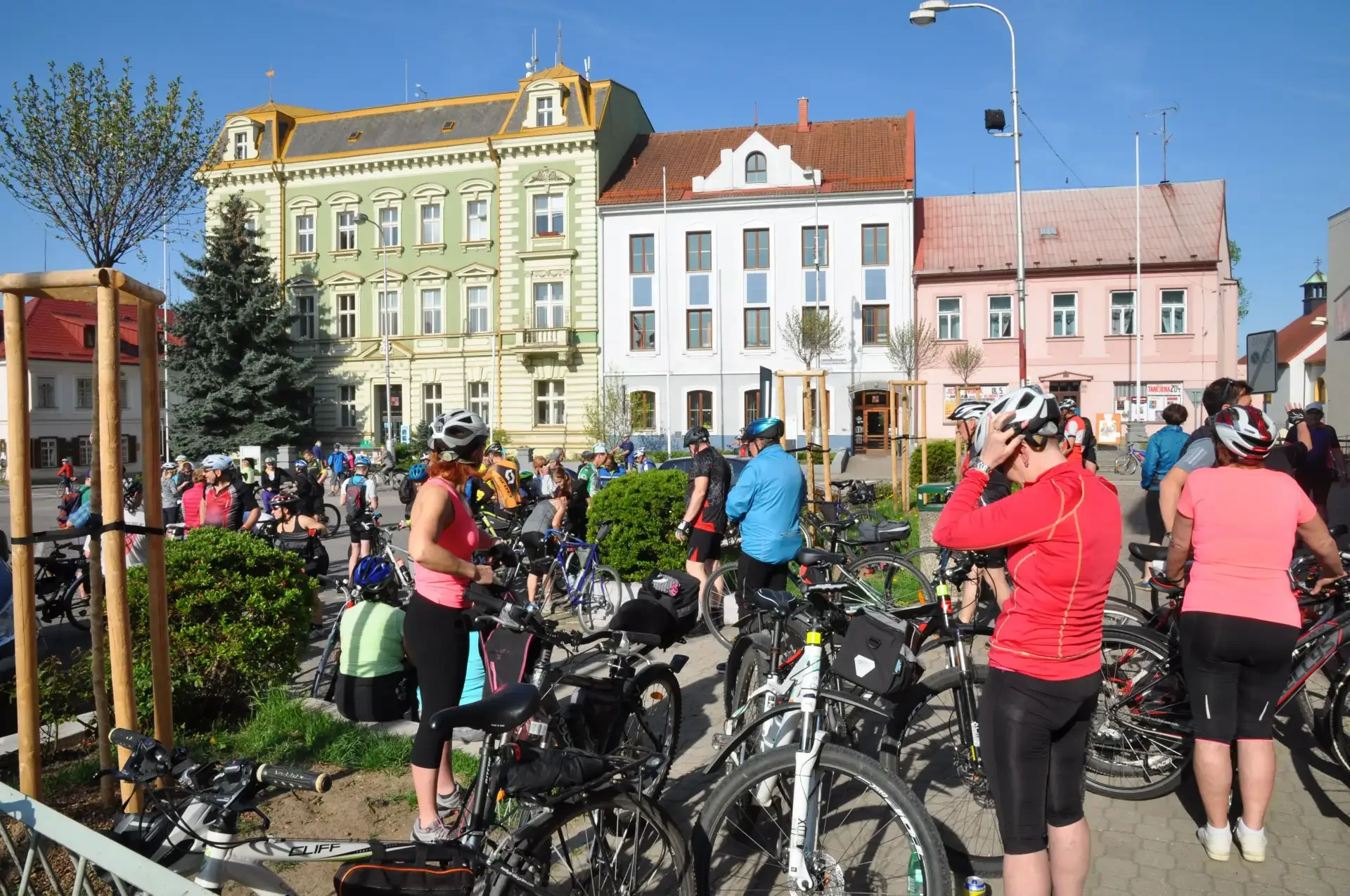 Cyklisté s koly a helmami se shromažďují na náměstí před barevnými domy