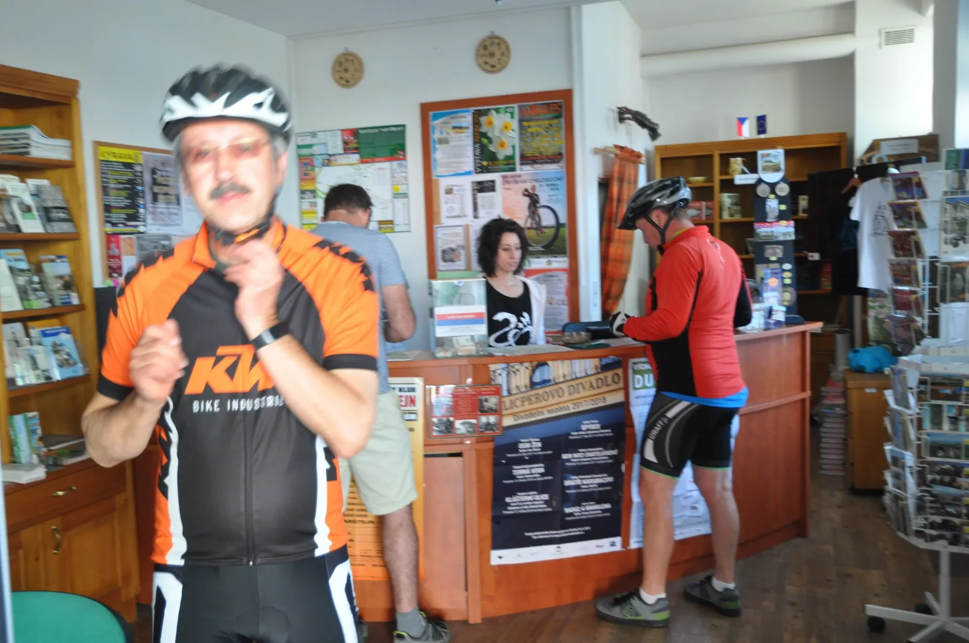 Cyklista v helmě a oranžovo-černém dresu v informačním centru s plakáty na zdi