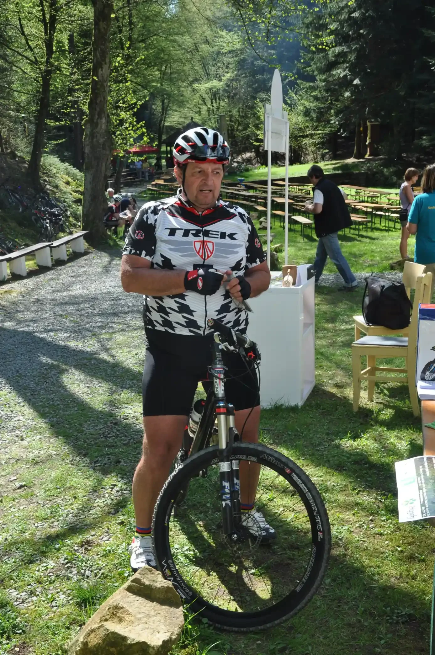 Cyklista v dresu Trek s horským kolem na trávníku u venkovních stolů