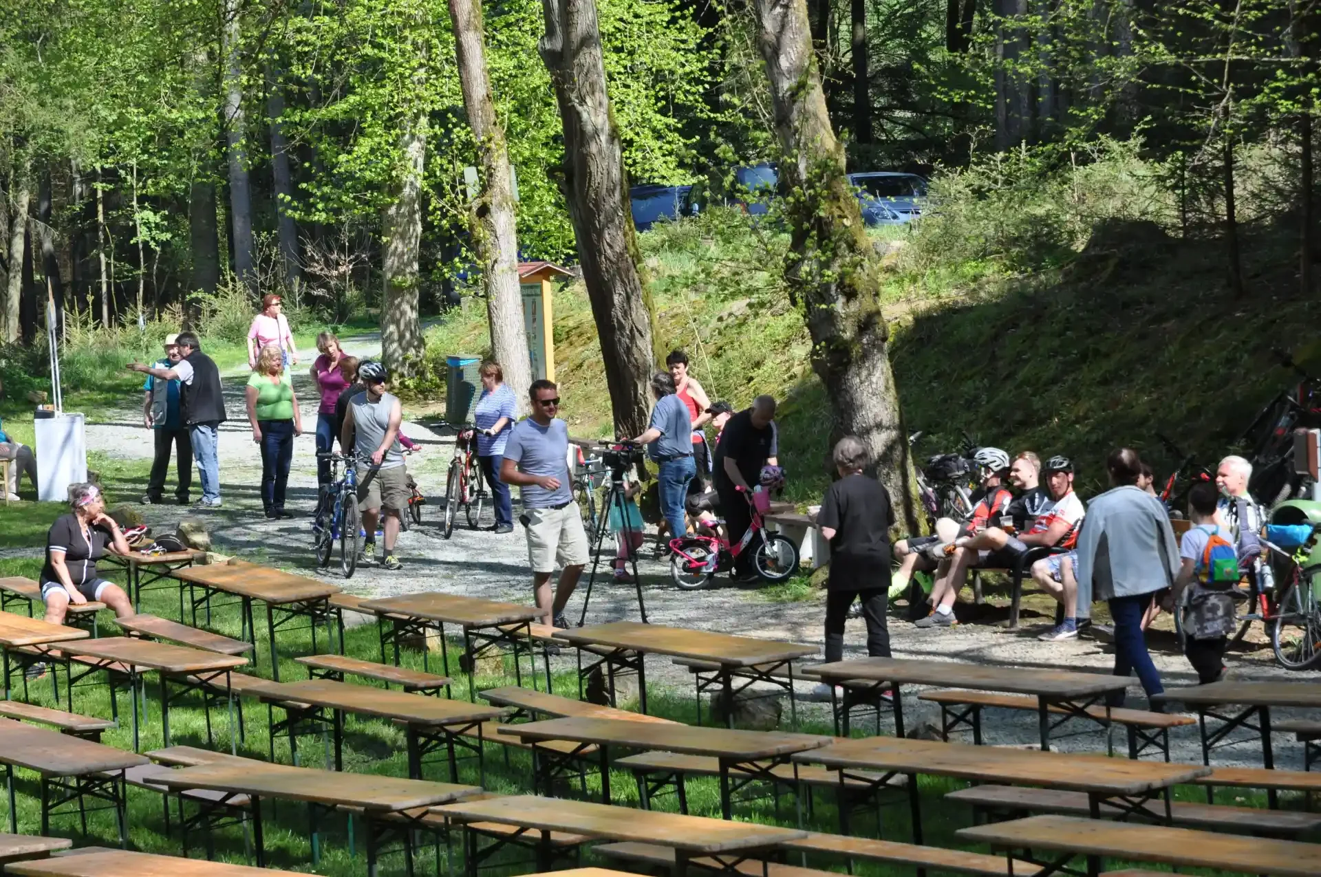 Návštěvníci u prázdných stolů v lese, v pozadí natáčení kamerou a zaparkovaná kola