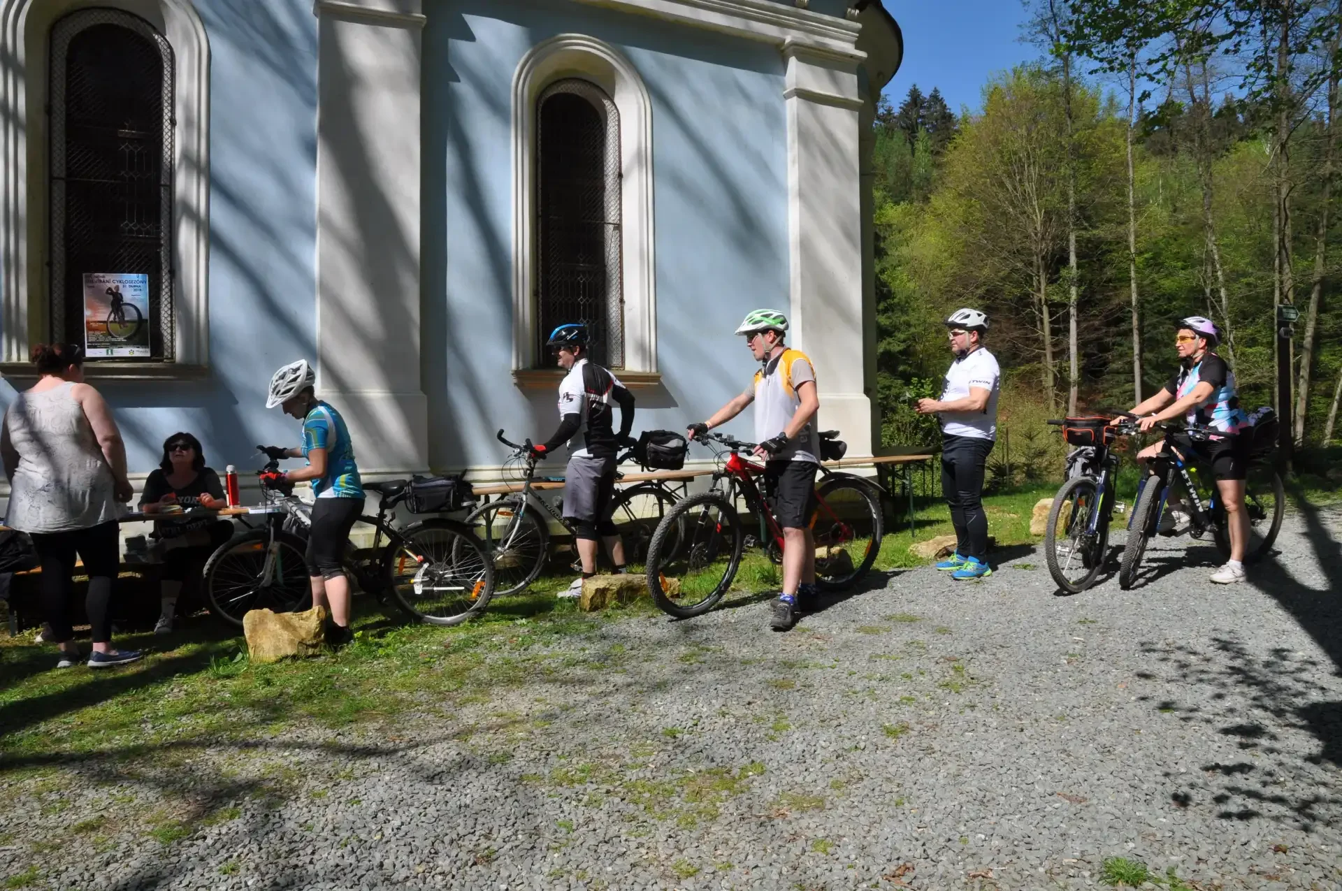 Cyklisté s koly stojí před bíle-modrou barokní kaplí u lesa