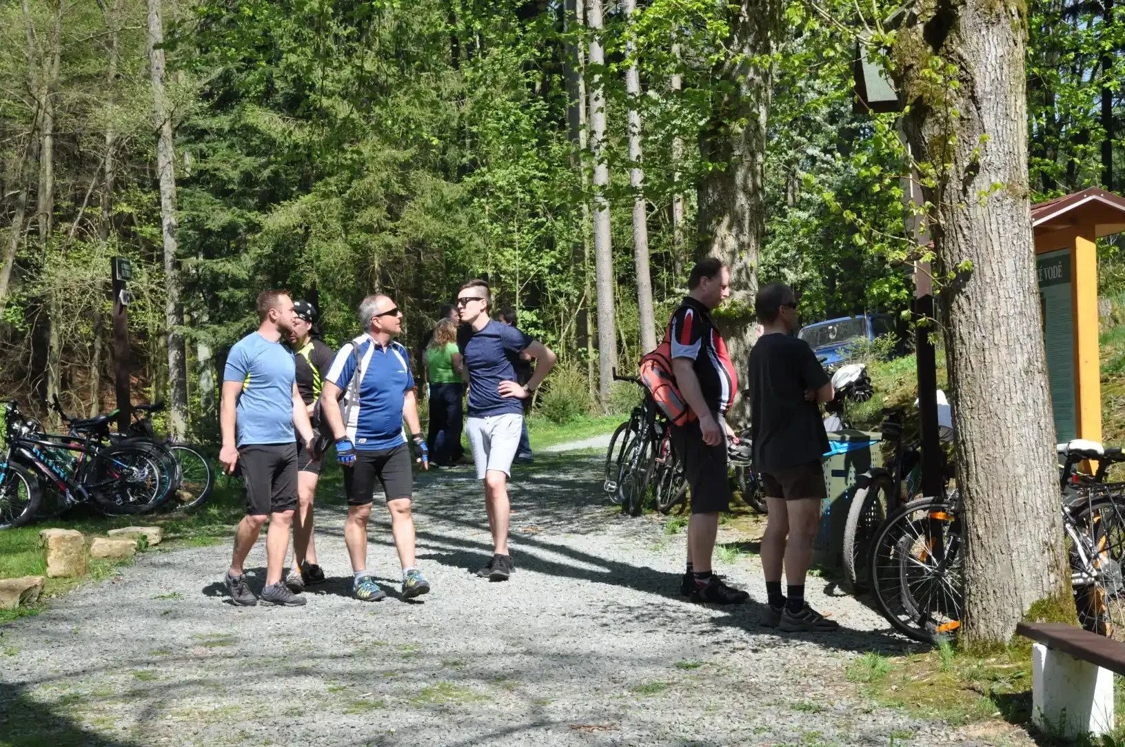 Skupina mužů diskutuje u lesní informační tabule, v pozadí zaparkovaná kola