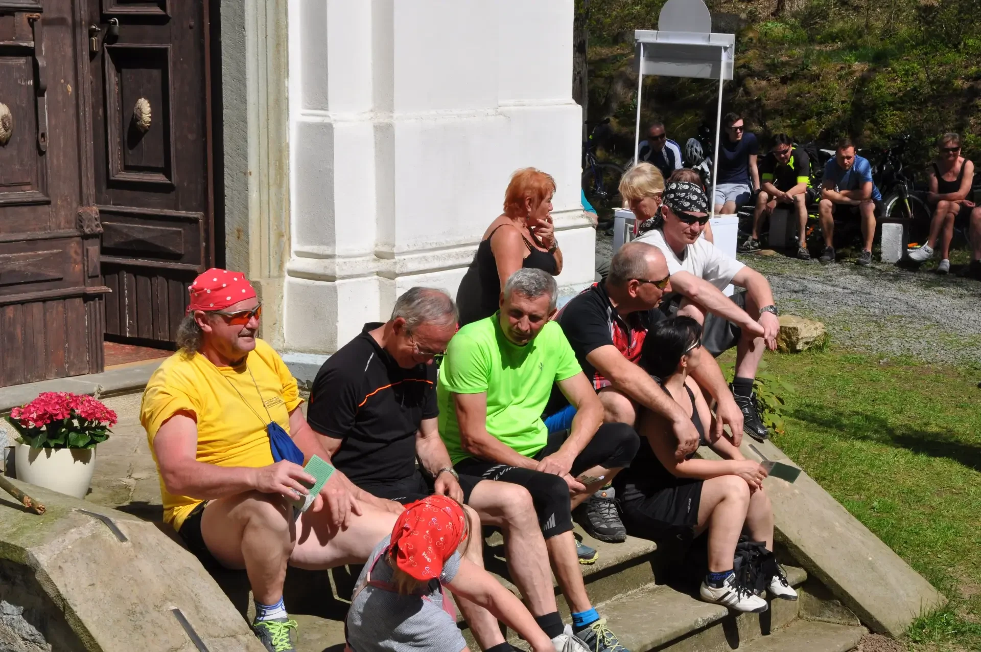 Skupina cyklistů odpočívá na schodech kaple na sluníčku