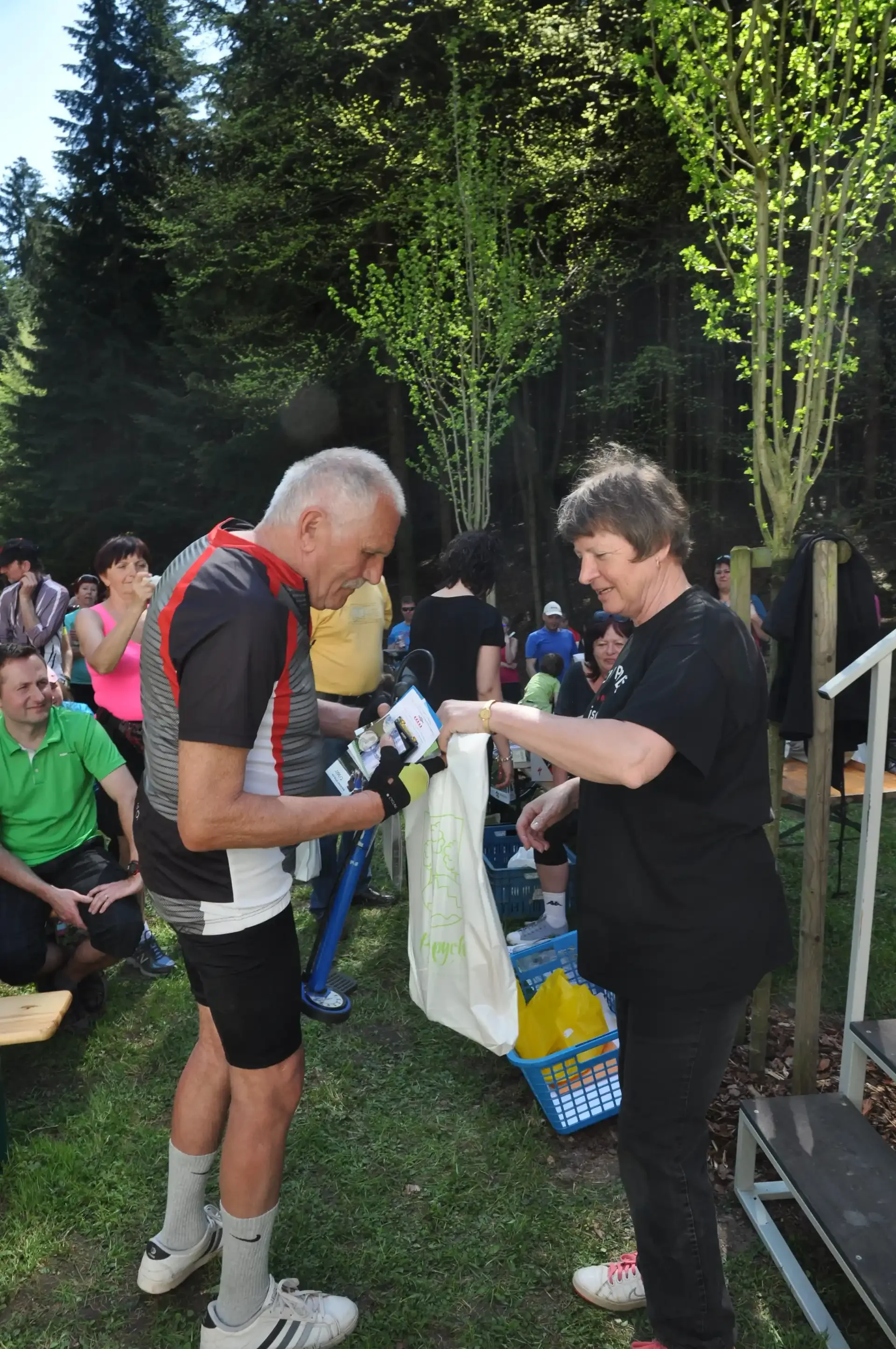 Cyklista si prohlíží obsah dárkové tašky, žena mu pomáhá, kolem přihlížející