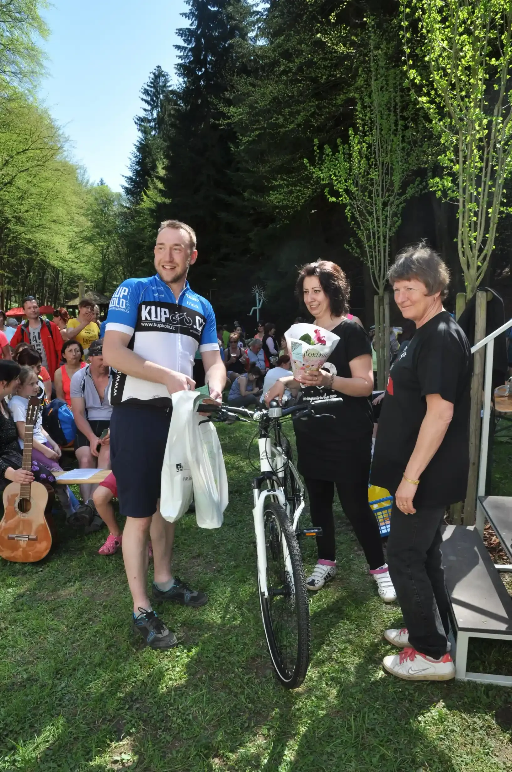 Cyklista v dresu Kup kolo.cz předává kolo a dárkovou tašku ženám s kyticí