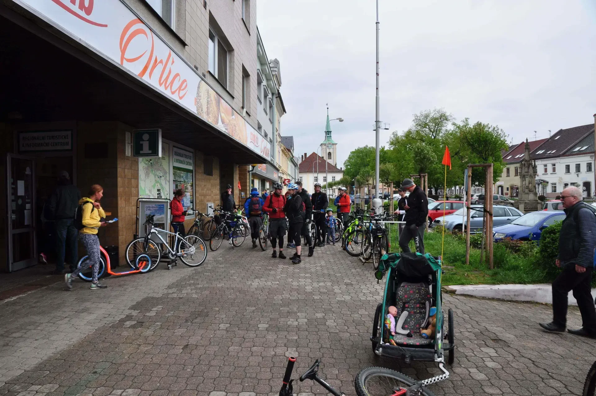 Skupina cyklistů s helmami a koly u budovy Regionálního turistického a informačního centra na náměstí, vpravo dětský vozík za kolem