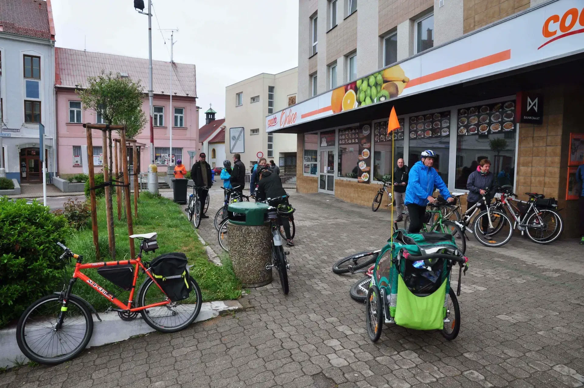 Cyklisté s koly a dětským vozíkem odpočívají na chodníku před obchodem Coop na náměstí ve městě
