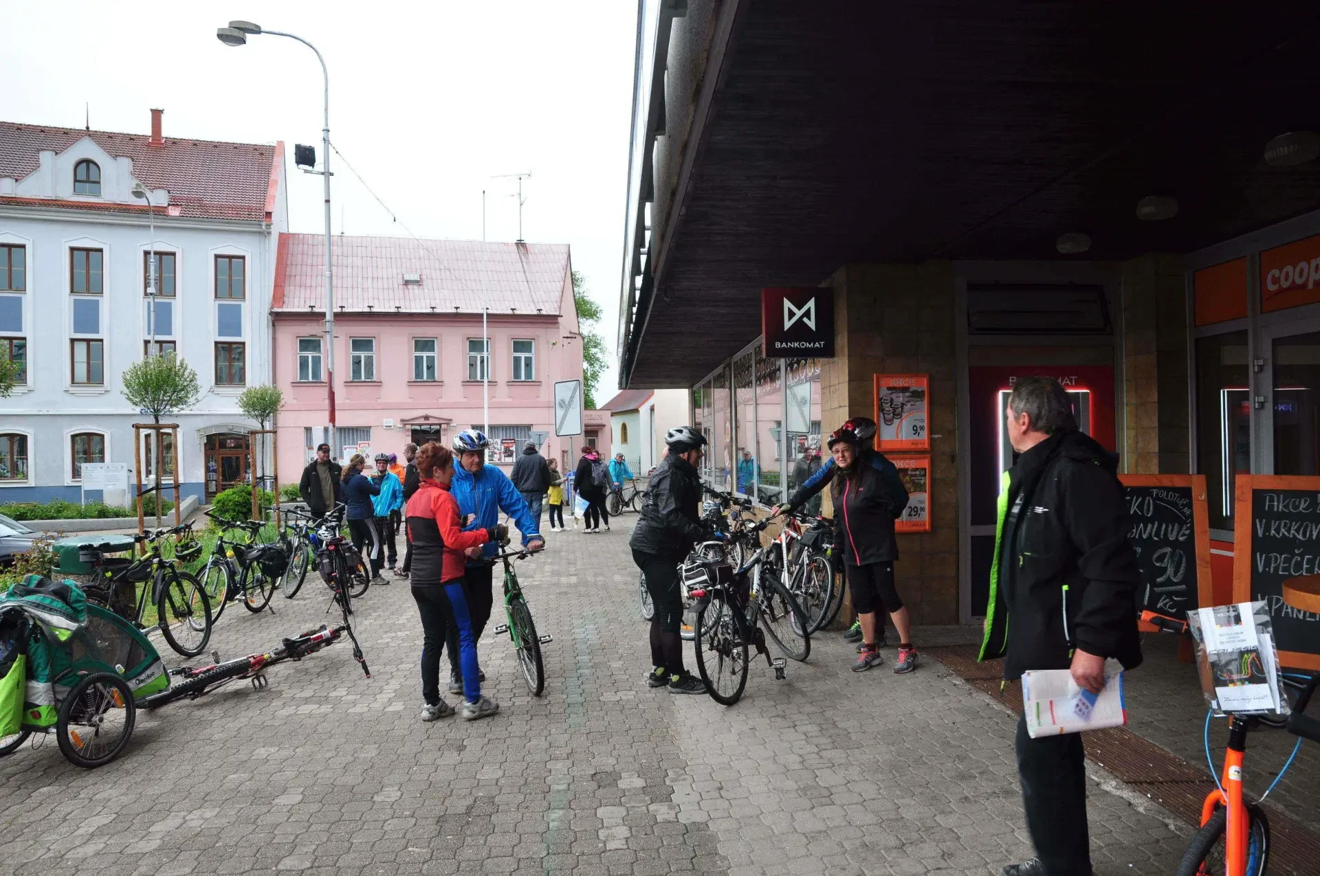 Cyklisté s koly se shromažďují na chodníku před restaurací a bankomatem na náměstí, v pozadí historické domy