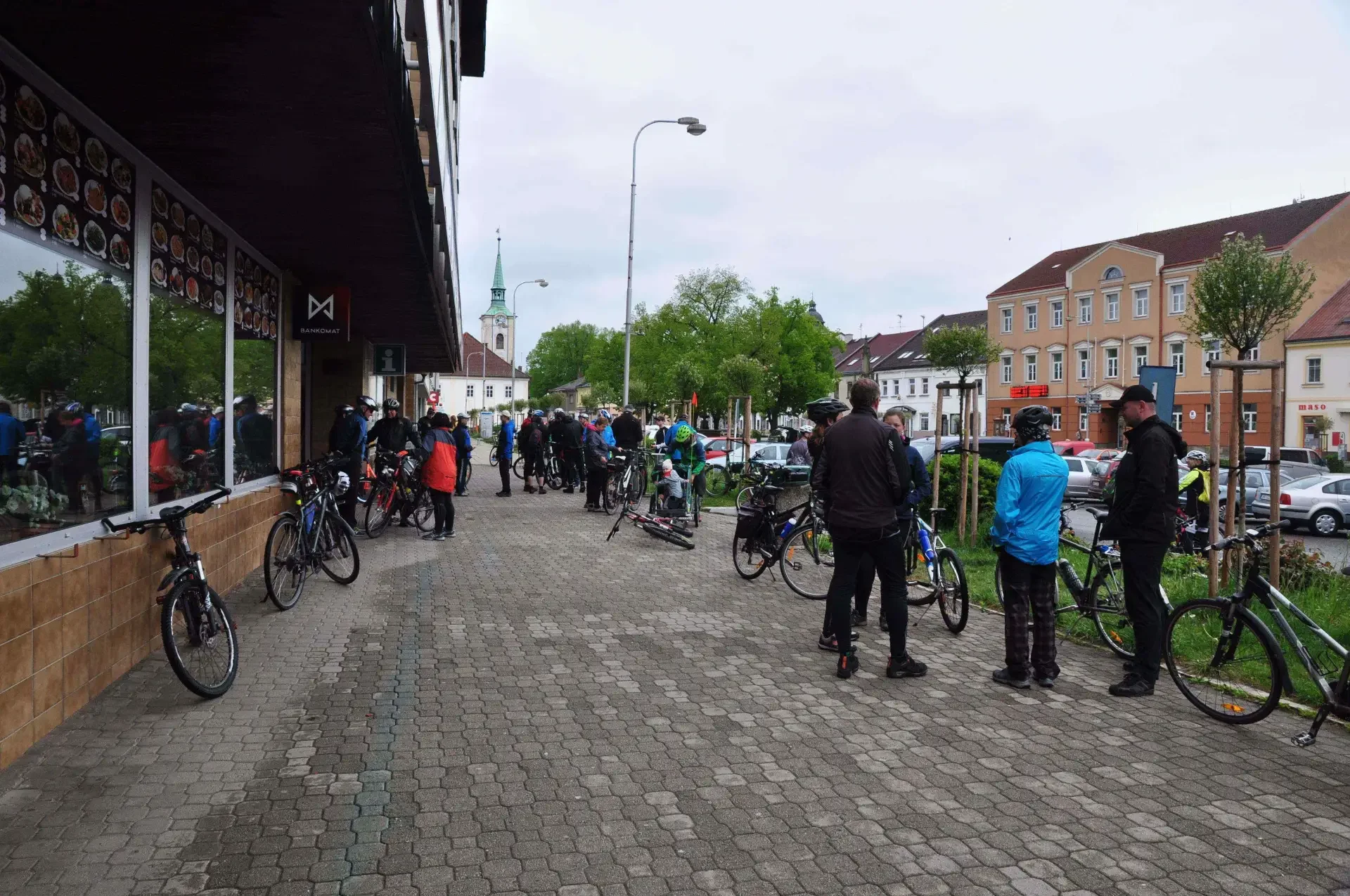 Skupina cyklistů s koly stojí na chodníku před restaurací, v pozadí kostelní věž a náměstí s domy