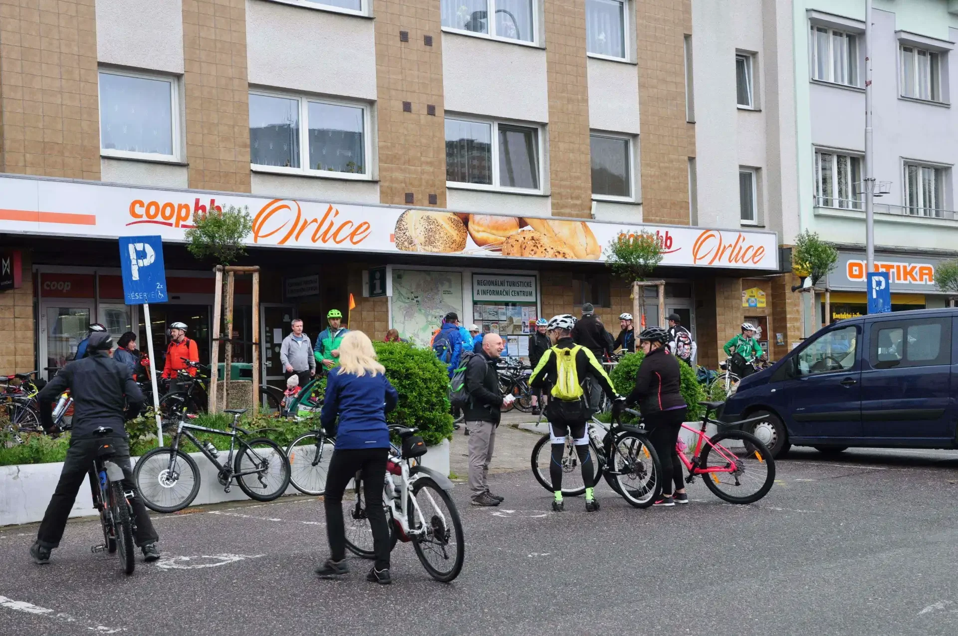 Cyklisté s koly se shromažďují před obchodem Coop na náměstí, v popředí dětský vozík za kolem