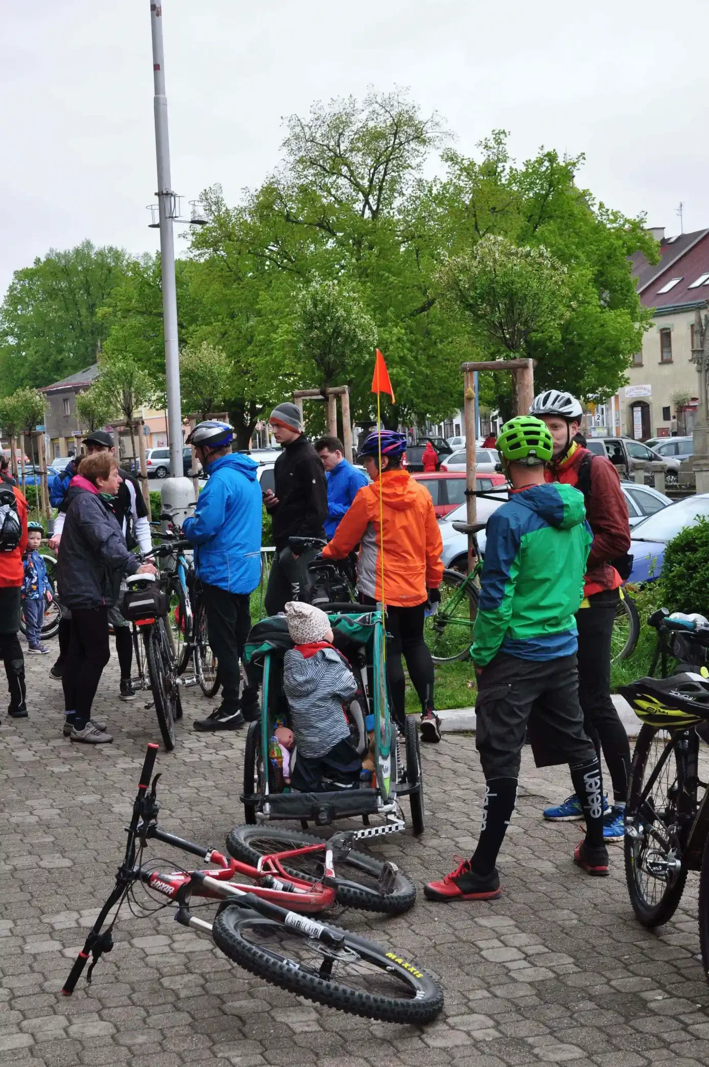 Skupina cyklistů s helmami a dítětem v přívěsném vozíku stojí na náměstí obklopená koly, v pozadí historické domy se sochou
