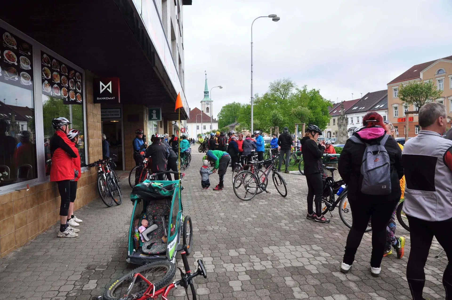 Cyklisté s koly stojí na chodníku před restaurací s bankomatem, v pozadí náměstí s kostelní věží a dětský přívěsný vozík v popředí