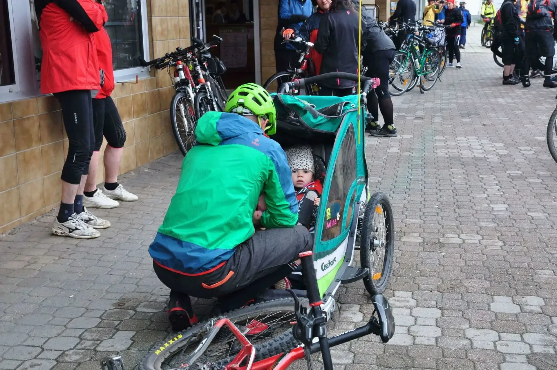 Muž v zelené bundě a cyklistické helmě si hraje s malým dítětem v přívěsném vozíku za kolem na chodníku před obchodem