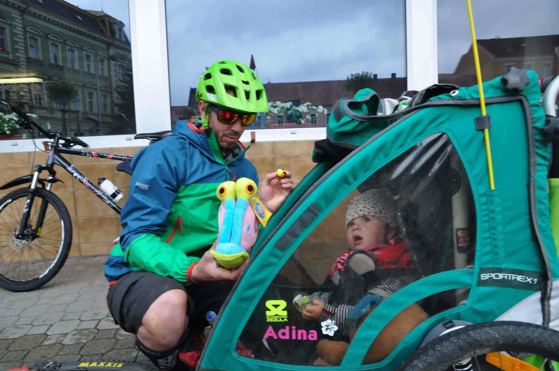 Muž v cyklistické helmě ukazuje plyšovou hračku dítěti sedícímu v zeleném přívěsném vozíku za kolem, v odrazu skla vidět náměstí