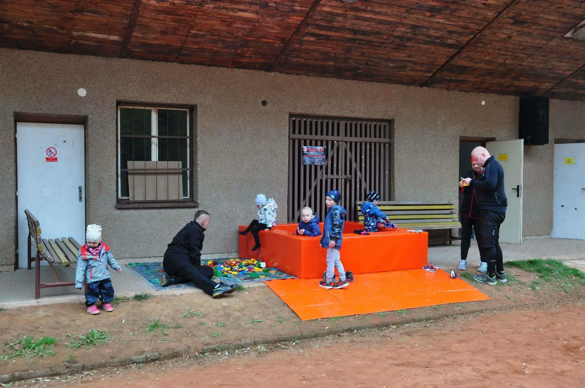 Velká skupina lidí u dlouhých stolů na travnaté ploše sportovního areálu, v pozadí zaparkovaná auta a dětské hřiště.