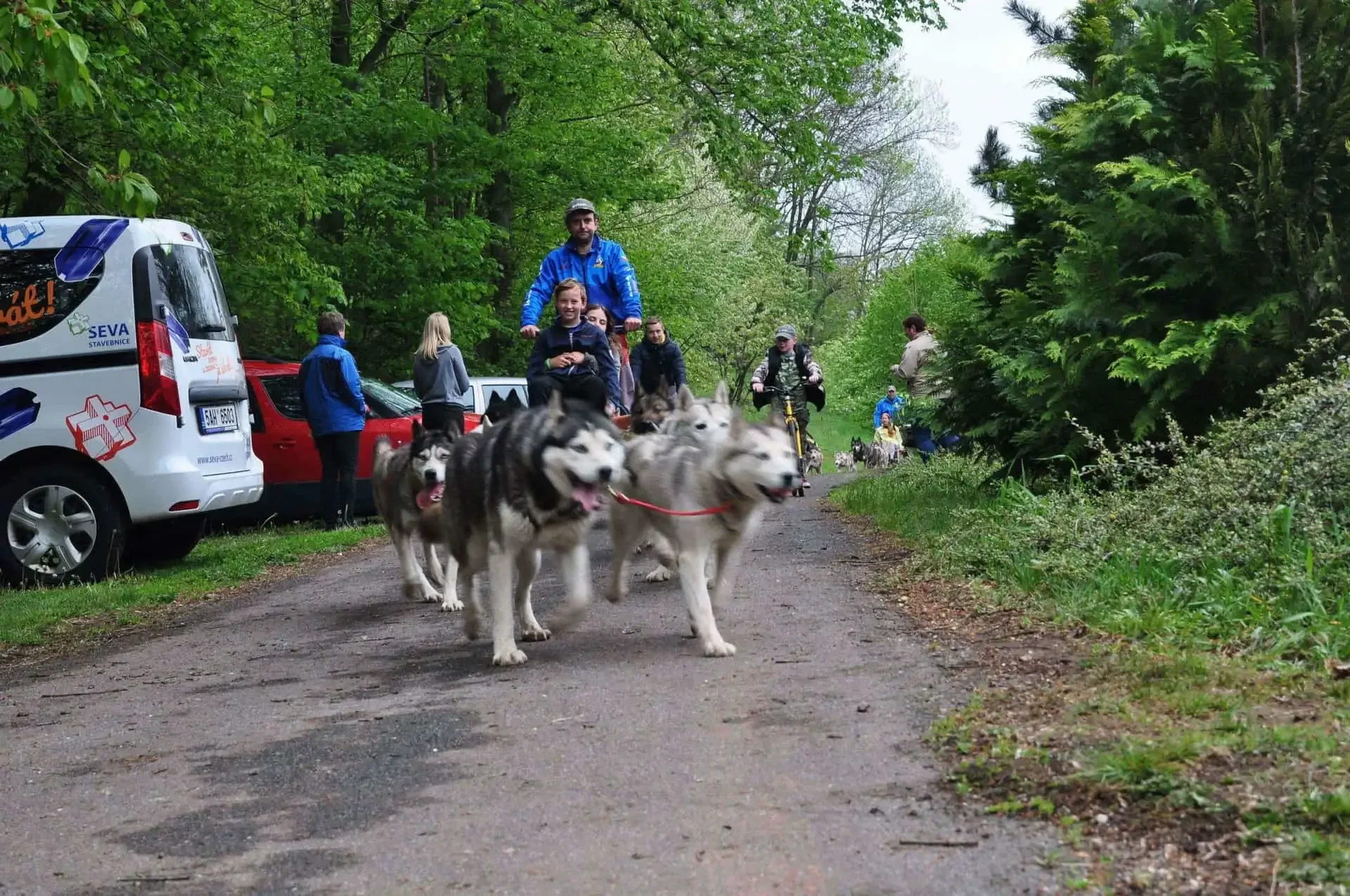 Spřežení huskyů táhne vozík s mužem a chlapcem po lesní cestě, vedle jedou na kolech další účastníci, v pozadí dodávka SEVA a přihlížející.