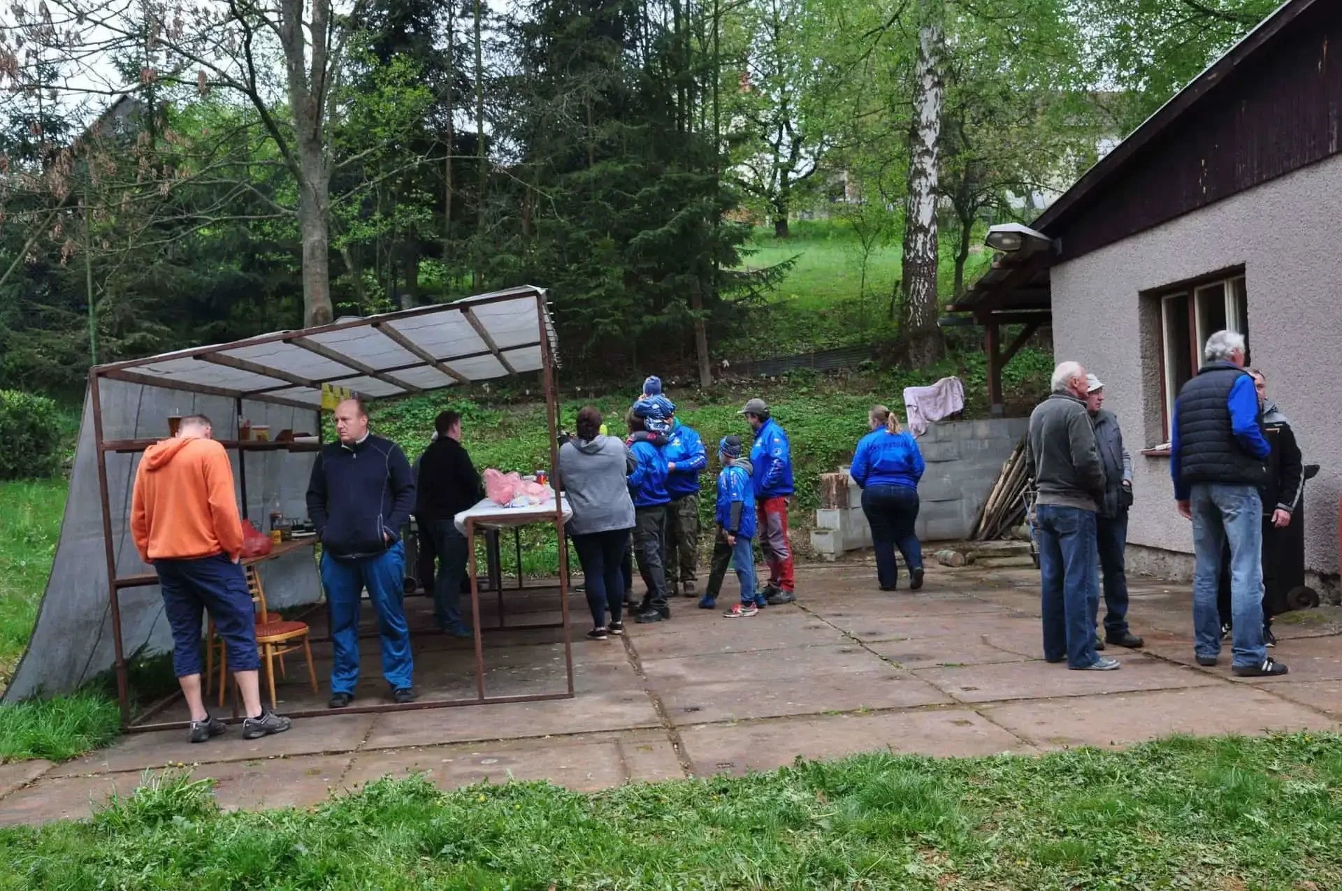 Skupina lidí u stánku s občerstvením a stolem na venkovní ploše před chatou, v pozadí zelený svah a stromy