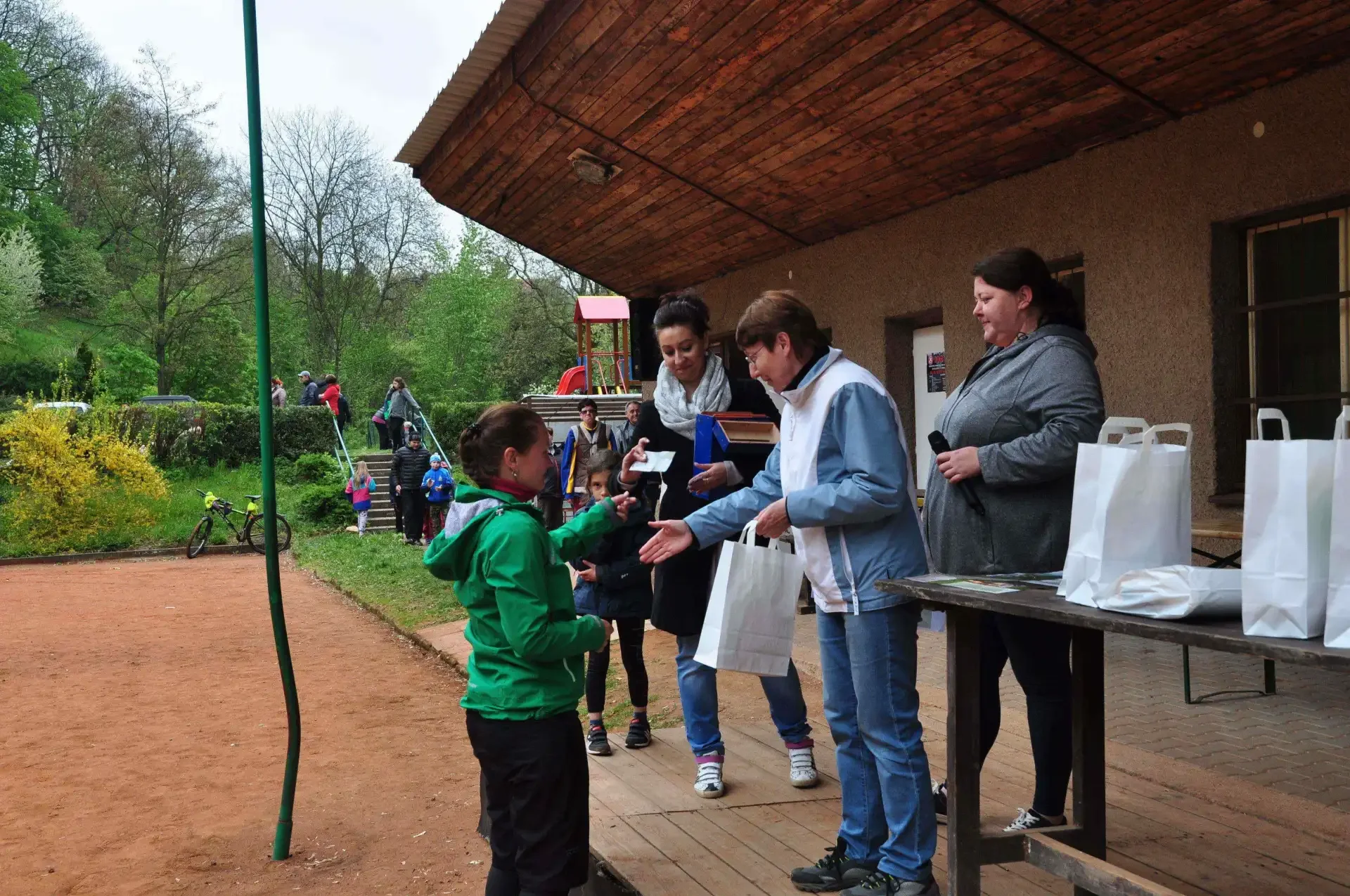 Žena v zelené bundě odchází od stolu s dárkovými taškami, v pozadí čeká dívka a další lidé na předávání cen před chatou