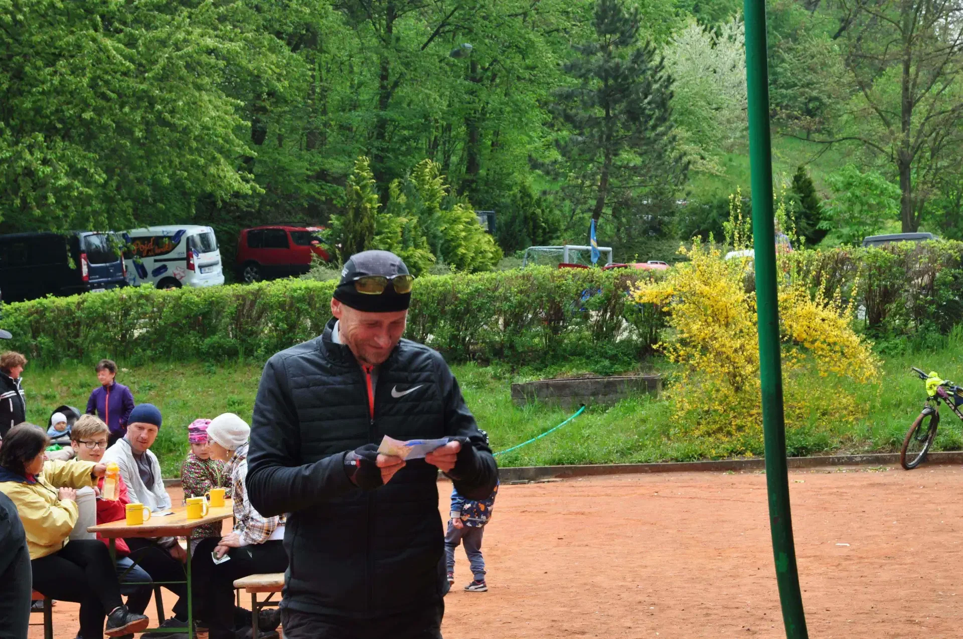 Muž v černé bundě, kšiltovce a slunečních brýlích se usmívá nad papírem s výsledky, v pozadí rodina u stolu a zaparkovaná auta.
