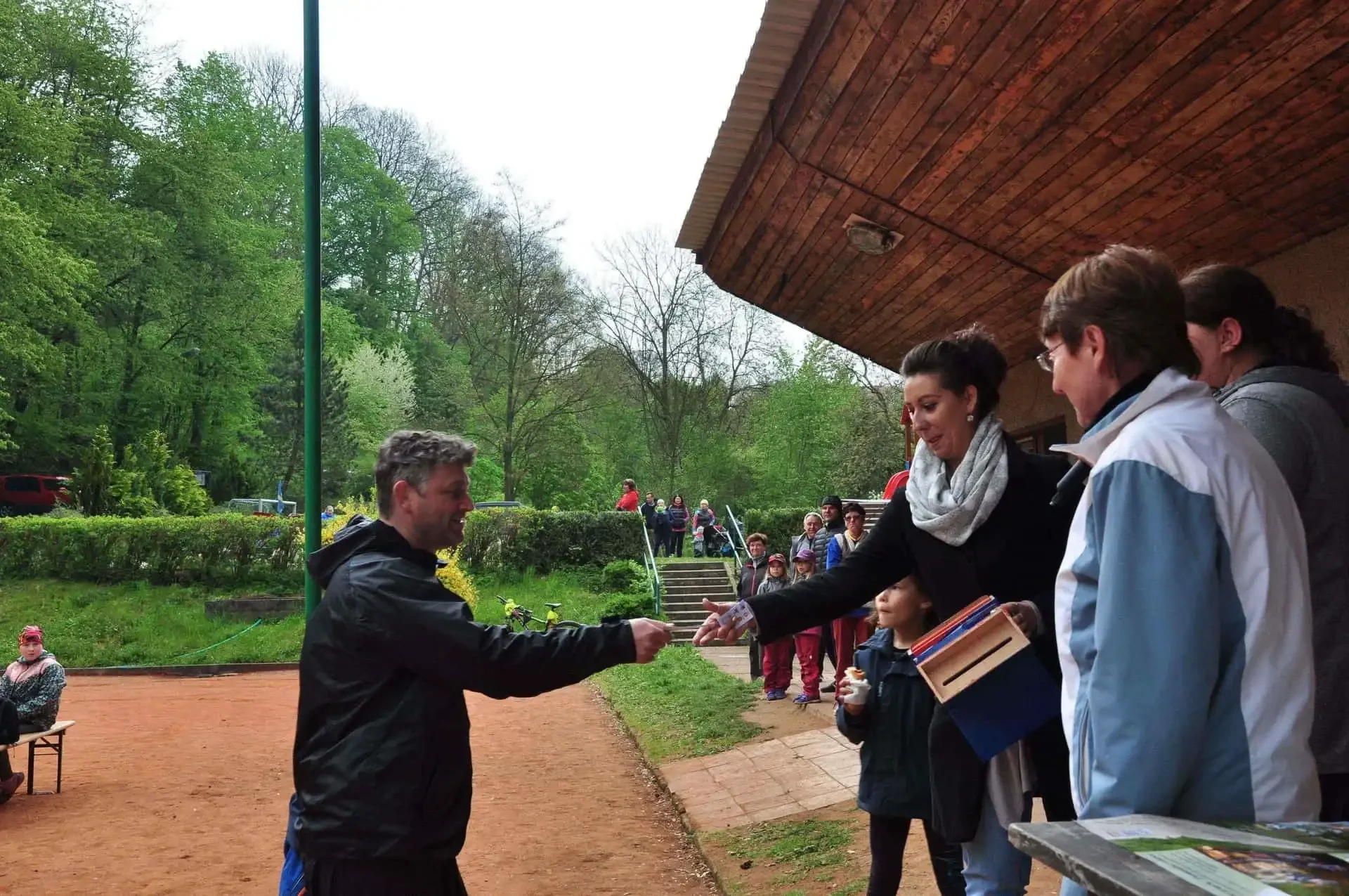 Muž se v předklonu s úsměvem loučí a podává ruku ženě v červené bundě s čelenkou, v pozadí hřiště a běžící chlapec.