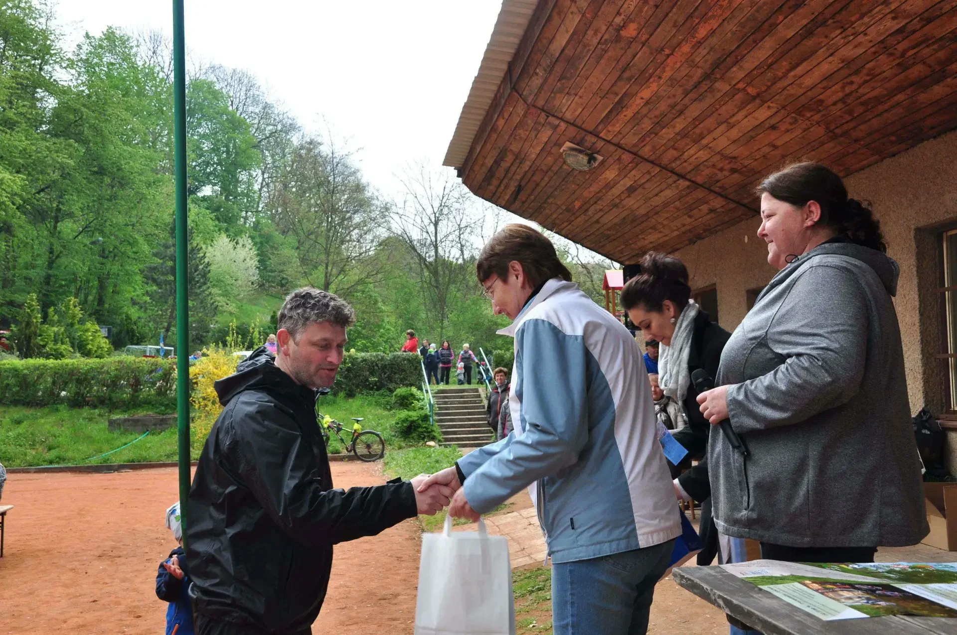 Žena v černobílé sportovní bundě si podává ruku se ženou v modrobílé bundě a přebírá dárkovou tašku, vpravo žena s mikrofonem.