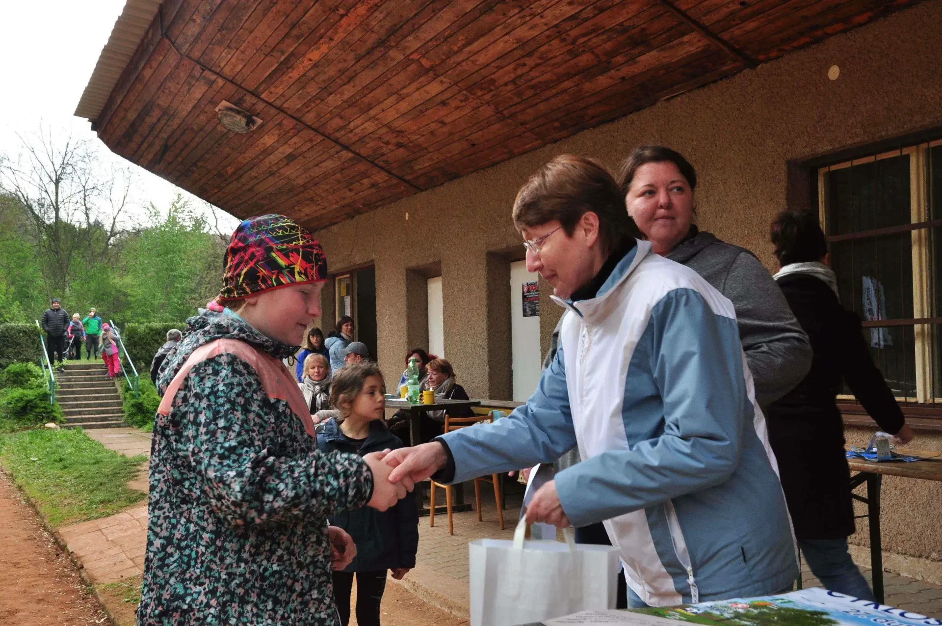 Dívka v barevném šátku si podává ruku se ženou a přebírá dárkovou tašku, kolem sedí lidé u stolu před klubovnou.