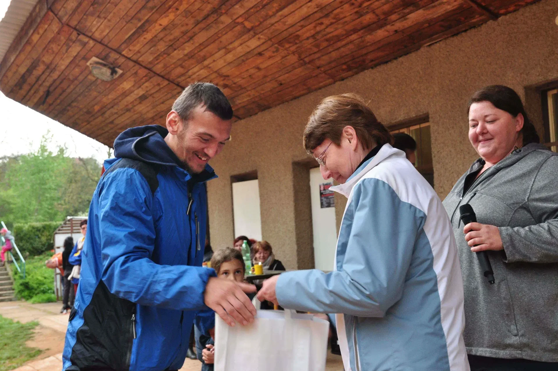 Muž v modré bundě si podává ruku se ženou v modrobílé bundě a přebírá zabalený dárek, vedle stojí dívka se šátkem a papíry.