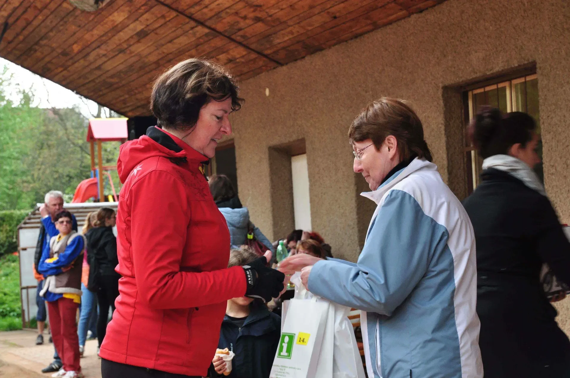 Žena v červené bundě s čelenkou poklekává a přebírá bílou dárkovou tašku od ženy v modrobílé bundě, v pozadí hřiště a běžící chlapec.