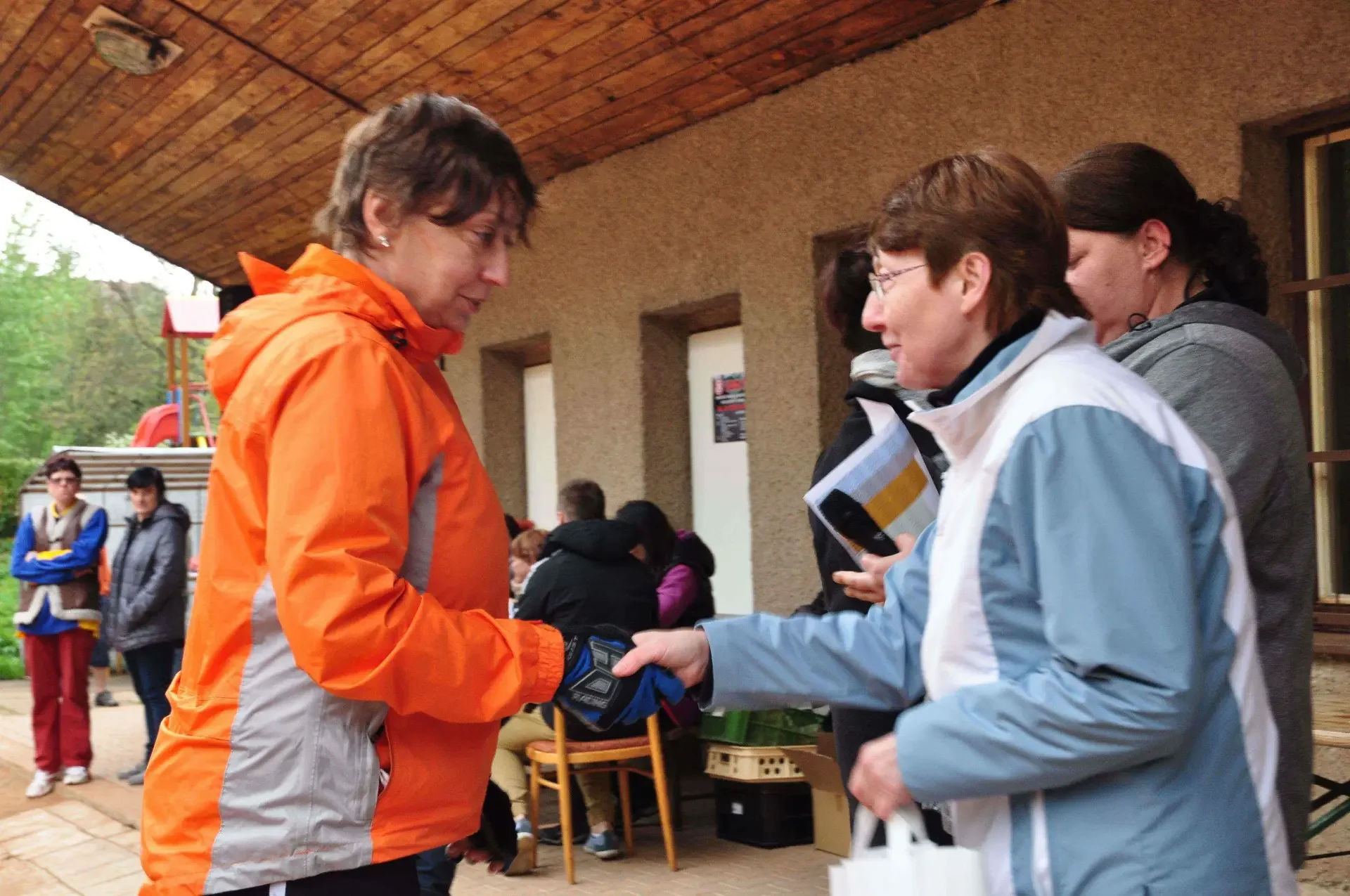Žena v oranžové bundě si podává ruku s další ženou v modrobílé bundě před dřevěným přístřeškem, kolem stojí přihlížející