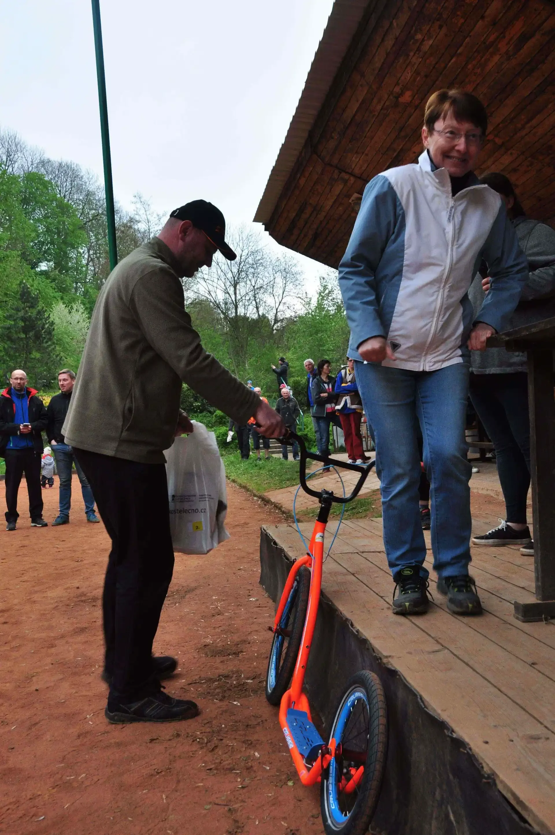 Muž předává výherci oranžovou koloběžku, žena vedle se dívá a usmívá, kolem přihlížejí další účastníci akce.