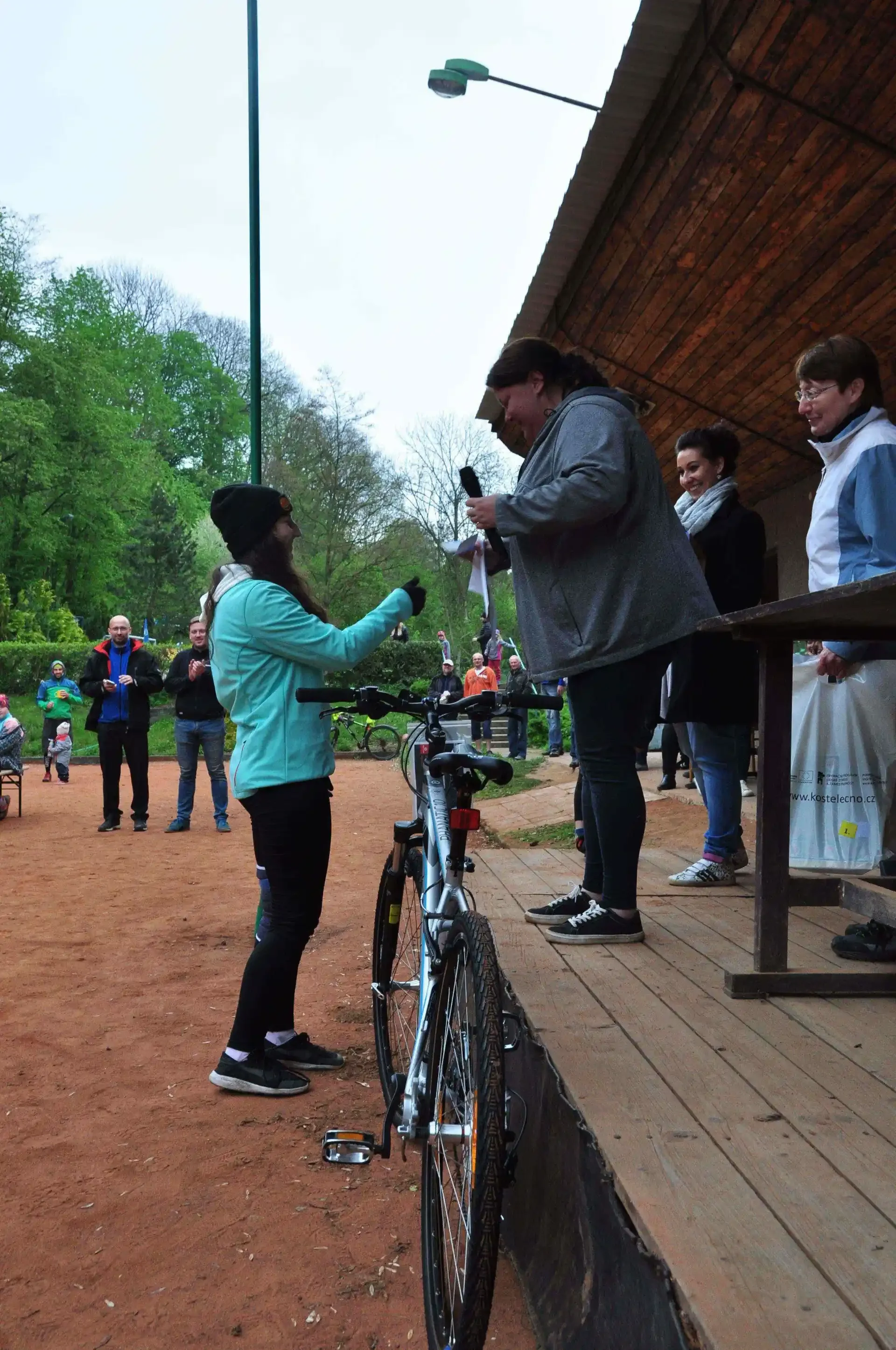 Žena v tyrkysové bundě s kolem po boku si povídá se ženou na dřevěné tribuně, v pozadí přihlížející lidé.