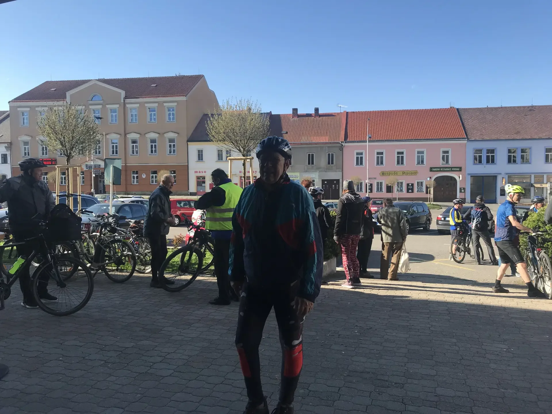 Muž v cyklistické helmě stojí na náměstí, v pozadí skupina cyklistů