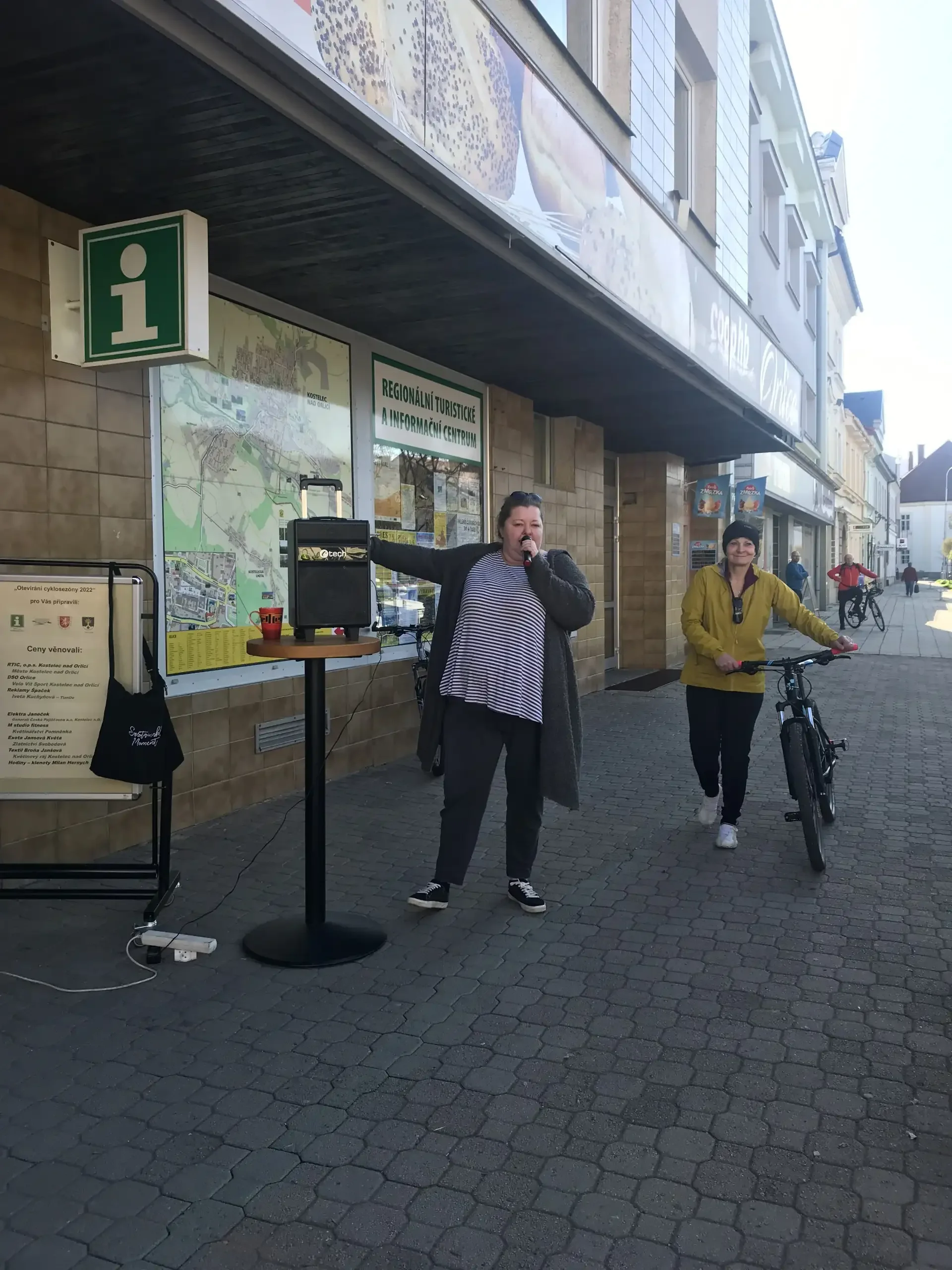 Žena mluví do mikrofonu před informačním centrem, kolem projíždí cyklistka