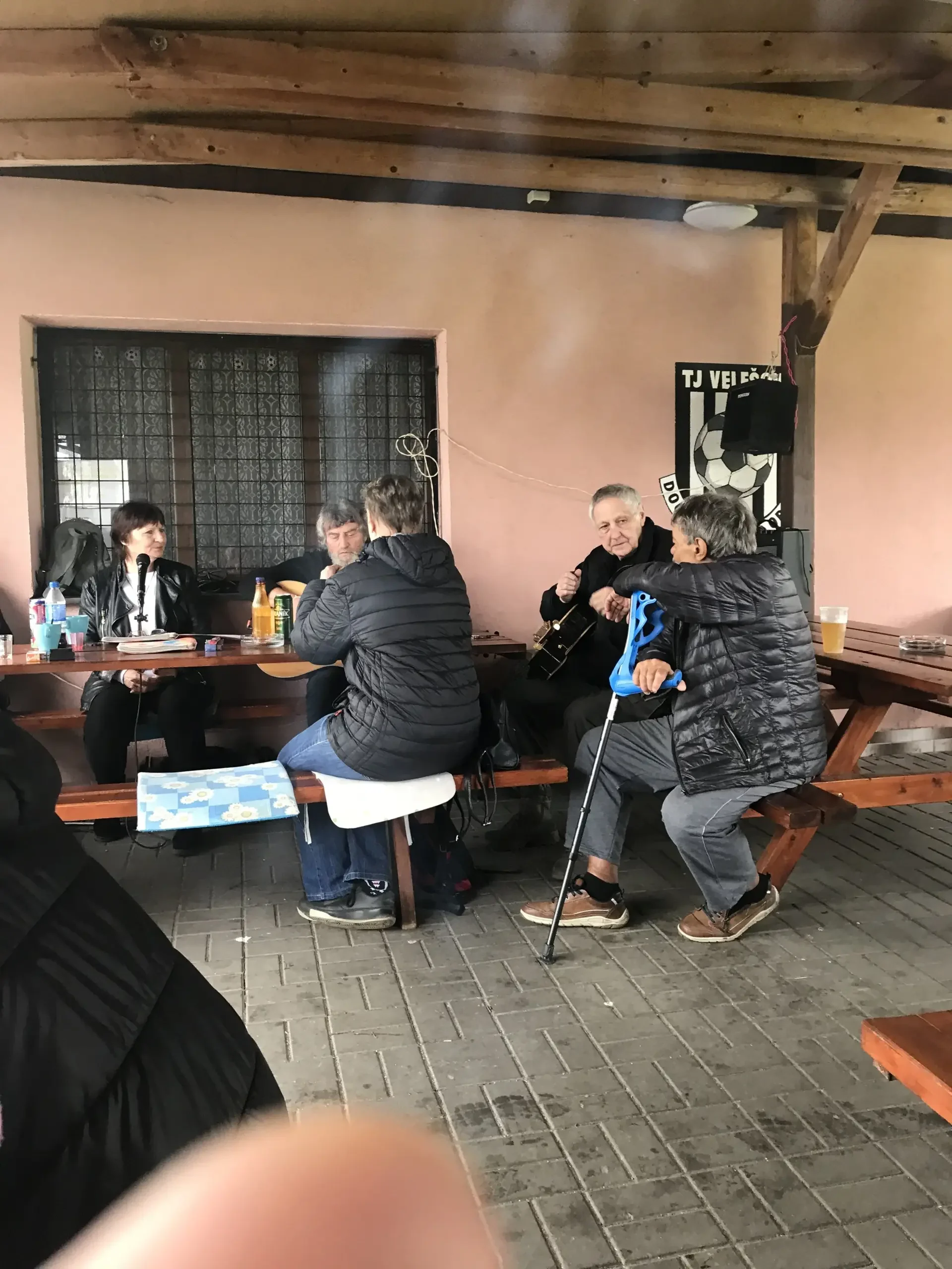 Hudebníci hrají na kytary pod přístřeškem, kolem sedí lidé