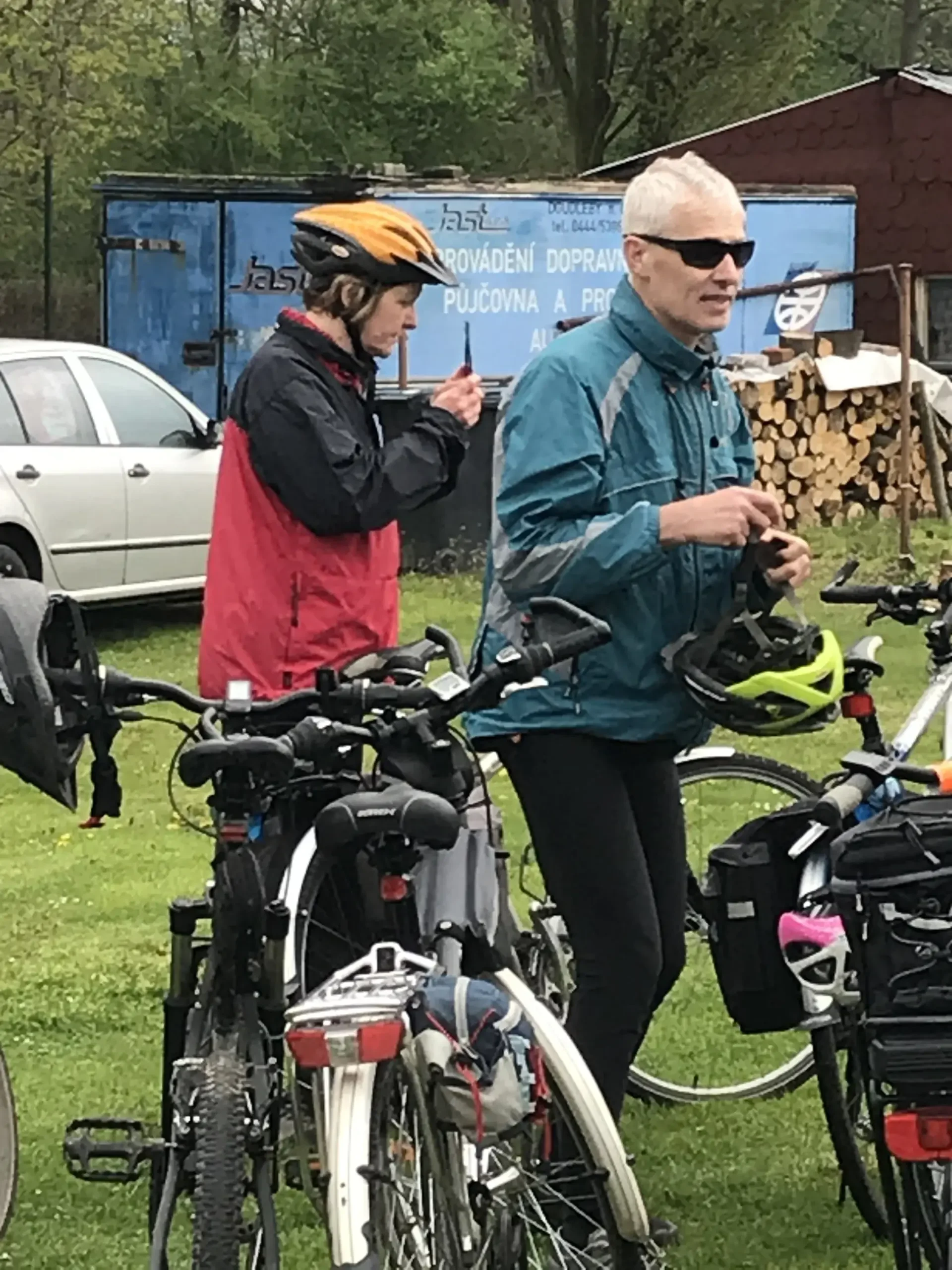 Dva cyklisté v pláštěnkách u kol na trávě před dřevnicí