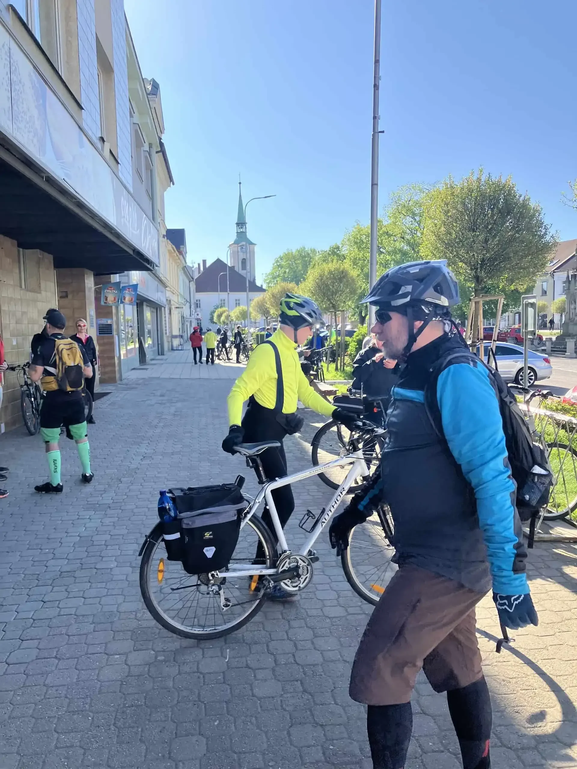 Cyklisté s koly na chodníku, v pozadí kostelní věž