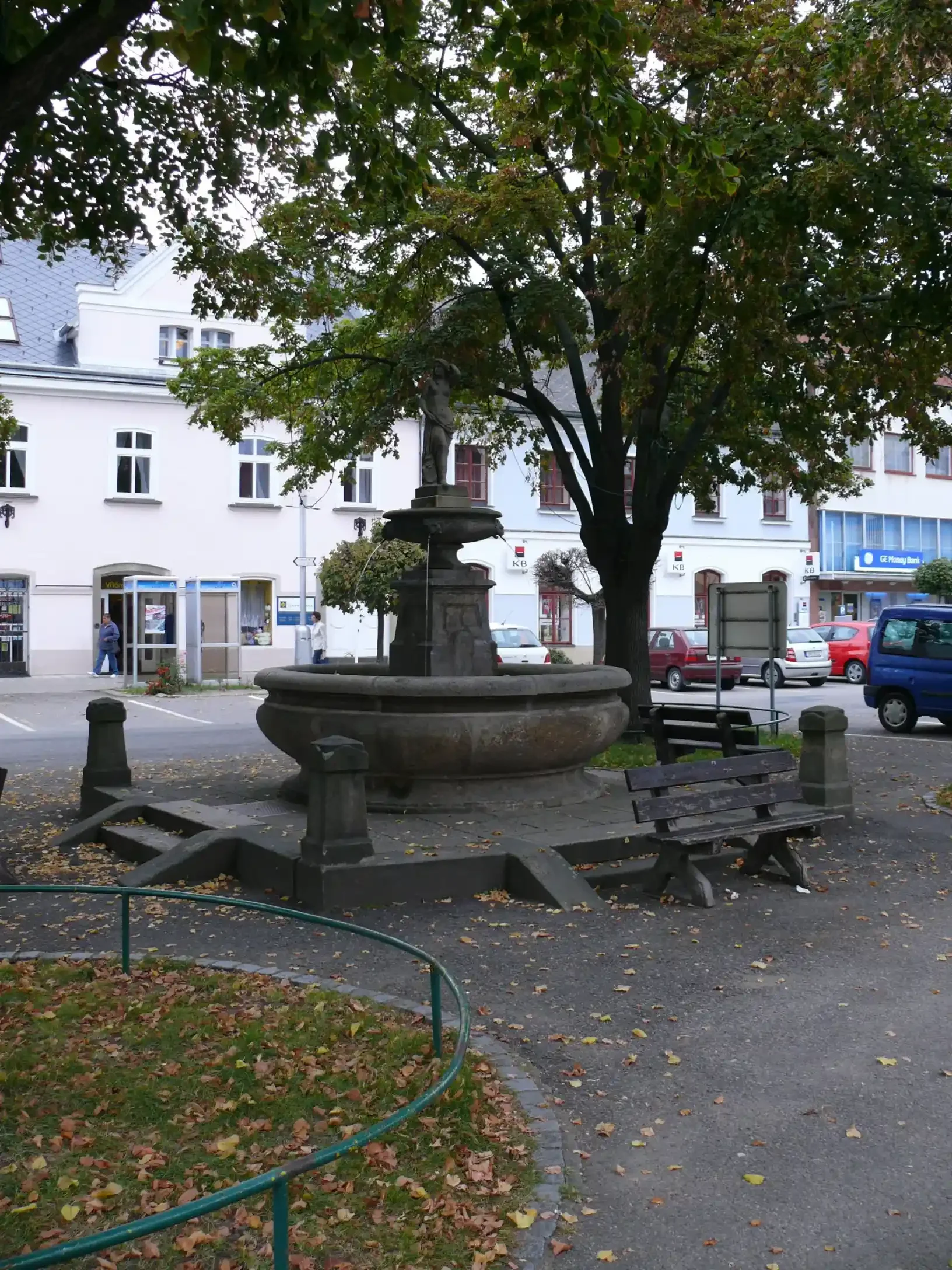 Kamenná kašna se sochou pod stromem na náměstí