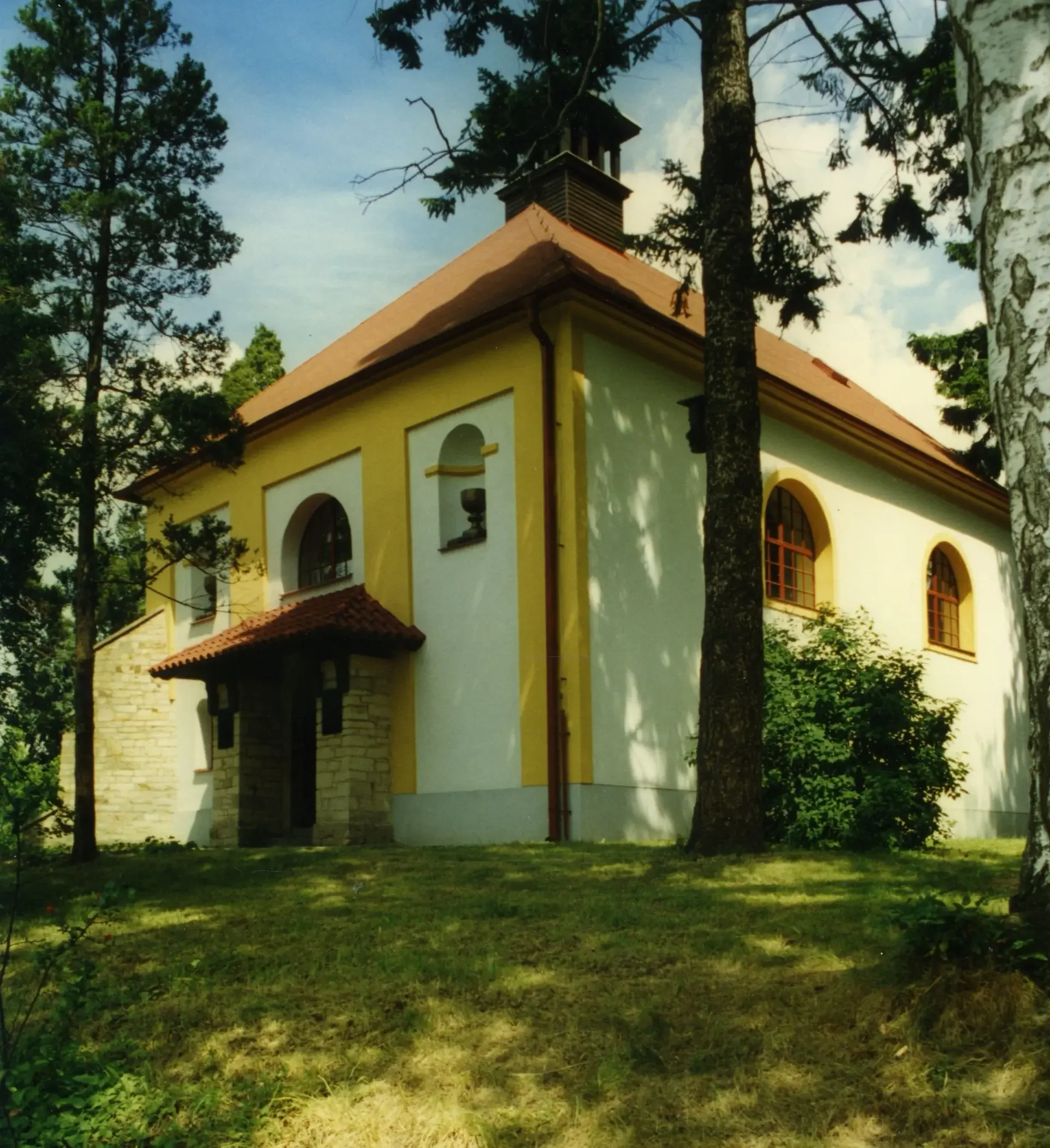 Žlutá kaplička s červenou střechou mezi stromy