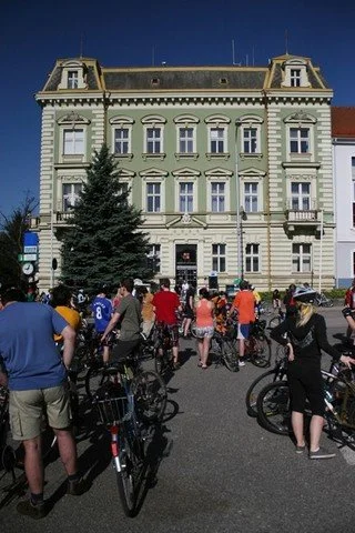 Cyklisté s koly se shromažďují před historickou budovou na náměstí