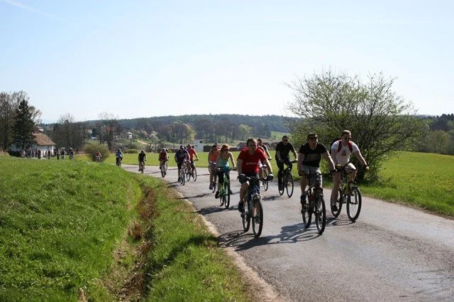 Skupina cyklistů jede po silnici mezi poli a zelení krajinou